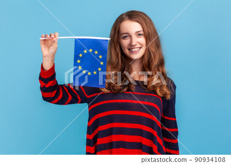Optimistic woman wearing striped casual style sweater, showing flag of European union, looking at camera with smile, EU association and community. Indoor studio shot isolated on blue background. Optimistic woman wearing striped casual style sweater, showing flag of European union, looking at camera with smile, EU association and community. Indoor studio shot isolated on blue background. 90934108