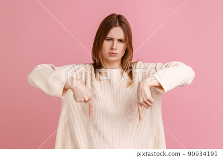 Portrait of strict bossy blond woman showing place down beside her, giving order with strict bossy expression, wearing white sweater. Indoor studio shot isolated on pink background. 90934191