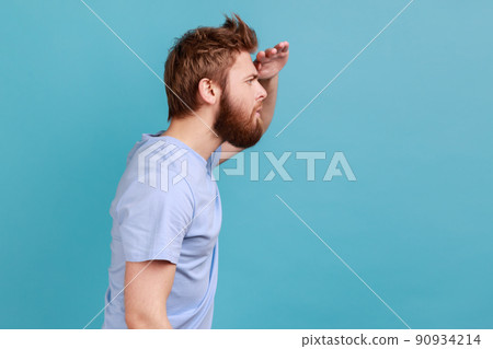 Side view of serious concentrated bearded man looking far away at distance with hand over head, attentively searching for bright future. Indoor studio shot isolated on blue background. 90934214