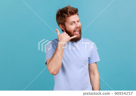 Give me a call. Portrait of positive optimistic bearded man smiling at camera and winking eye, making call me gesture with fingers, flirting. Indoor studio shot isolated on blue background. Give me a call. Portrait of positive optimistic bearded man smiling at camera and winking eye, making call me gesture with fingers, flirting. Indoor studio shot isolated on blue background. 90934257