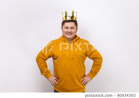 Man egoistically looking at camera and smiling, posing with crown on head, pretending to be king, privileged status, wearing urban style hoodie. Indoor studio shot isolated on white background. 90934303