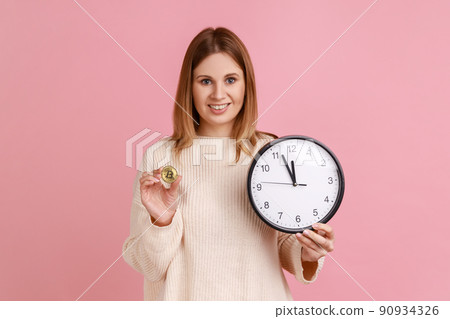 Portrait of positive rich blond woman holding golden coin of crypto currency and wall clock, time is money, expressing positive, wearing white sweater. Indoor studio shot isolated on pink background. Portrait of positive rich blond woman holding golden coin of crypto currency and wall clock, time is money, expressing positive, wearing white sweater. Indoor studio shot isolated on pink background. 90934326