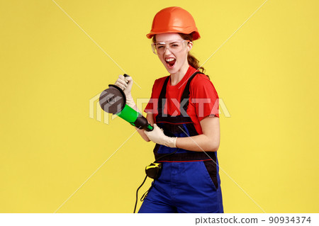 Portrait of crazy carpenter woman working with angle grinder, having excited facial expressing, screaming, wearing overalls and protective helmet. Indoor studio shot isolated on yellow background. 90934374