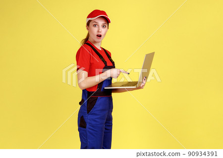 Portrait of shocked astonished worker woman working online on laptop computer, pointing at notebook display with open mouth, wearing overalls. Indoor studio shot isolated on yellow background. 90934391