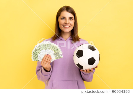 Portrait of woman with toothy smile holding soccer ball and fan of euro, celebrating victory on football championship, betting, wearing purple hoodie. Indoor studio shot isolated on yellow background. Portrait of woman with toothy smile holding soccer ball and fan of euro, celebrating victory on football championship, betting, wearing purple hoodie. Indoor studio shot isolated on yellow background. 90934393