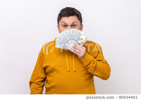 Portrait of dark haired middle aged man covering half of face with many dollar banknotes, looking at camera, wearing urban style hoodie. Indoor studio shot isolated on white background. Portrait of dark haired middle aged man covering half of face with many dollar banknotes, looking at camera, wearing urban style hoodie. Indoor studio shot isolated on white background. 90934401