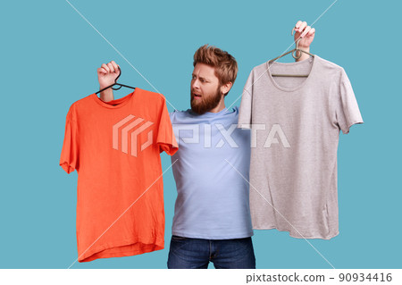 Portrait of confused bearded man with frowning face holding two hangers with gray and orange T-shirts, hard to choose the best attire. Indoor studio shot isolated on blue background. Portrait of confused bearded man with frowning face holding two hangers with gray and orange T-shirts, hard to choose the best attire. Indoor studio shot isolated on blue background. 90934416
