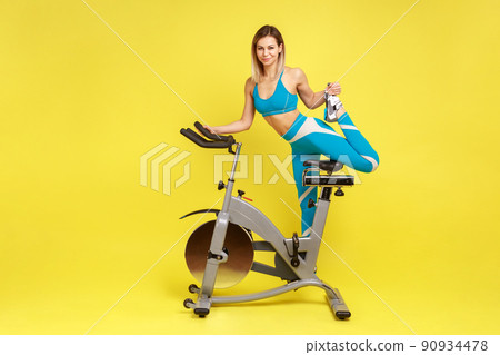 Full length portrait of slim woman with perfect body standing on one leg on exercise bike, looking at camera, wearing blue sportswear. Indoor studio shot isolated on yellow background. Full length portrait of slim woman with perfect body standing on one leg on exercise bike, looking at camera, wearing blue sportswear. Indoor studio shot isolated on yellow background. 90934478
