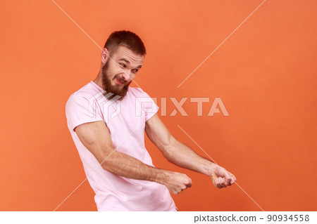 Portrait of strong bearded man keeping fists up, holding invisible virtual rope and pulling object, striving hard with effort, wearing pink T-shirt. Indoor studio shot isolated on orange background. 90934558