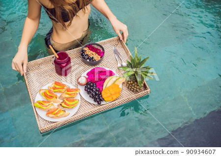 Breakfast tray in swimming pool, floating breakfast in luxury hotel. Girl relaxing in the pool drinking smoothies and eating fruit plate, smoothie bowl by the hotel pool. Exotic summer diet. Tropical Breakfast tray in swimming pool, floating breakfast in luxury hotel. Girl relaxing in the pool drinking smoothies and eating fruit plate, smoothie bowl by the hotel pool. Exotic summer diet. Tropical 90934607