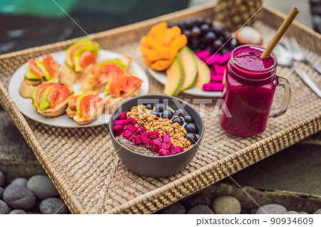 Breakfast on a tray with fruit, buns, avocado sandwiches, smoothie bowl by the pool. Summer healthy diet, vegan breakfast. Tasty vacation concept 90934609