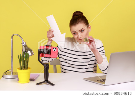 Sad upset woman office worker showing bitcoin and white arrow pointing down, sitting at workplace and recording video for her business blog. Indoor studio studio shot isolated on yellow background. Sad upset woman office worker showing bitcoin and white arrow pointing down, sitting at workplace and recording video for her business blog. Indoor studio studio shot isolated on yellow background. 90934613