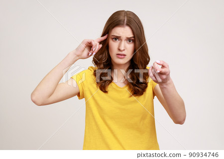 You are stupid. Self confident egoistic teenager girl wearing yellow T-shirt holding finger near temple and pointing on you, showing stupid gesture. Indoor studio shot isolated on gray background. You are stupid. Self confident egoistic teenager girl wearing yellow T-shirt holding finger near temple and pointing on you, showing stupid gesture. Indoor studio shot isolated on gray background. 90934746