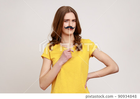 Portrait of funny childish teenager girl in yellow T-shirt holding fake mustache accessory over face, looking at camera, having festive good mood. Indoor studio shot isolated on gray background. Portrait of funny childish teenager girl in yellow T-shirt holding fake mustache accessory over face, looking at camera, having festive good mood. Indoor studio shot isolated on gray background. 90934848