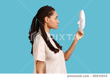 Side view of confident serious woman with dreadlocks holding and looking at white mask with attentive look, trying to understand hiding personality. Indoor studio shot isolated on blue background. 90934854