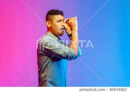 Side view portrait of handsome young adult man in shirt looking far away through monocular gesture, spying shocking content. Indoor studio shot isolated on colorful neon light background. Side view portrait of handsome young adult man in shirt looking far away through monocular gesture, spying shocking content. Indoor studio shot isolated on colorful neon light background. 90934862