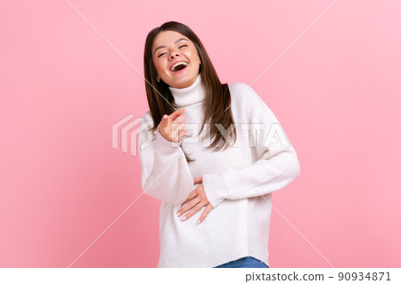 Happy female laughing out loud and pointing finger to camera, hearing funny joke, holding belly, wearing white casual style sweater. Indoor studio shot isolated on pink background. 90934871
