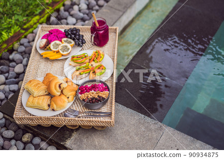 Breakfast on a tray with fruit, buns, avocado sandwiches, smoothie bowl by the pool. Summer healthy diet, vegan breakfast. Tasty vacation concept 90934875