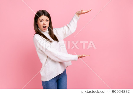 Woman presents copy space between hands, looks surprised, holds empty place for commercial idea, wearing white casual style sweater. Indoor studio shot isolated on pink background. 90934882
