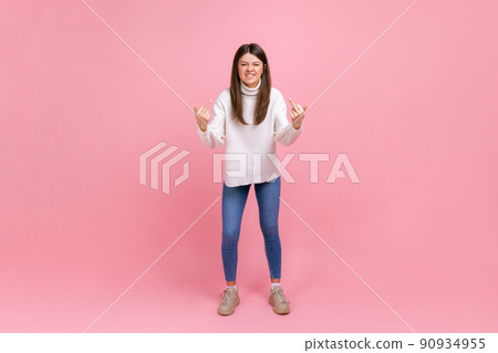 Full length portrait of aggressive girl standing and shows fuck sign with both hands into the camera, wearing white casual style sweater. Indoor studio shot isolated on pink background. 90934955