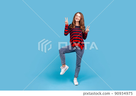 Full length portrait of happy woman wearing striped casual style sweater showing peace sign, v gesture and smiling, celebrating victory. Indoor studio shot isolated on blue background. Full length portrait of happy woman wearing striped casual style sweater showing peace sign, v gesture and smiling, celebrating victory. Indoor studio shot isolated on blue background. 90934975