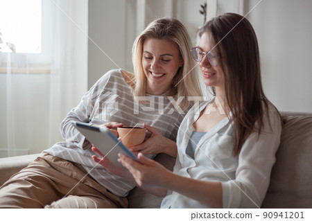 Two young women friends using tablet together, sitting on the couch smiling and fun Two young women friends using tablet together, sitting on the couch smiling and fun 90941201