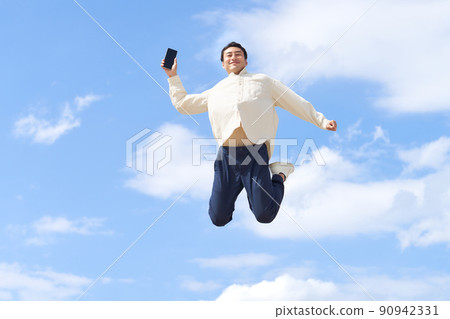 A young man jumping with a smartphone in his hand under the blue sky 90942331