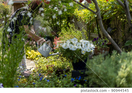 Senior woman watering plants with a white watering can in the garden of the house 90944545