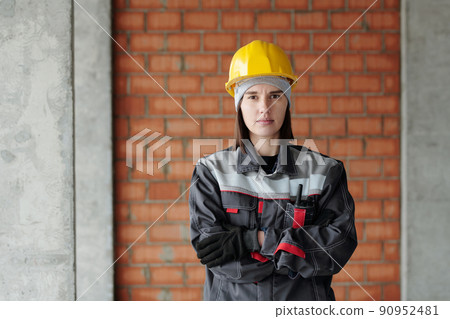 Young serious female engineer in protective helmet and workwear looking at you Young serious female engineer in protective helmet and workwear looking at you 90952481