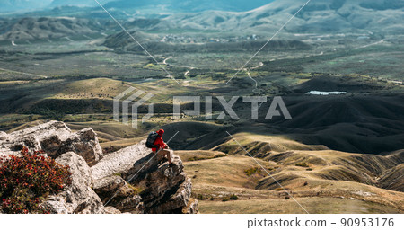A male traveler is sitting on the edge of a cliff. A male traveler is sitting on a rock with a bird's-eye view of the valley. The concept of recreation and tourism. The man enjoys the beautiful views A male traveler is sitting on the edge of a cliff. A male traveler is sitting on a rock with a bird's-eye view of the valley. The concept of recreation and tourism. The man enjoys the beautiful views 90953176