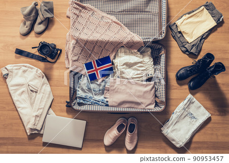 Woman's clothes, laptop, camera and flag of Iceland lying on the parquet floor near and in the open suitcase. Travel concept 90953457