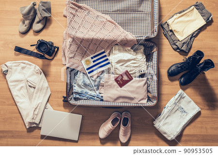 Woman's clothes, laptop, camera, russian passport and flag of Uruguay  lying on the parquet floor near and in the open suitcase. Travel concept 90953505
