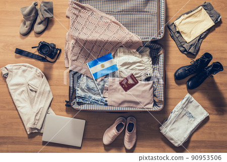 Woman's clothes, laptop, camera, russian passport and flag of Argentina  lying on the parquet floor near and in the open suitcase. Travel concept 90953506
