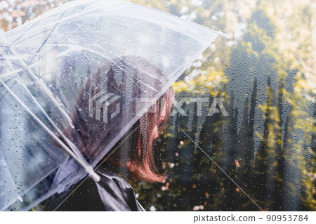 Autumn. Lonely unhappy woman walking in a park, garden with transparent umbrella at rain. View through a window with raindrops Autumn. Lonely unhappy woman walking in a park, garden with transparent umbrella at rain. View through a window with raindrops 90953784