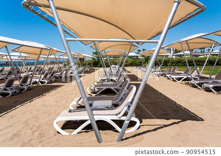 A rows of sunbeds at a deserted sandy beach in turkish resort. Sun loungers waiting for tourists in a hotel A rows of sunbeds at a deserted sandy beach in turkish resort. Sun loungers waiting for tourists in a hotel 90954515