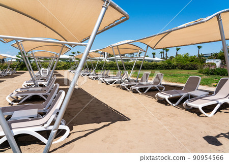 A rows of sunbeds at a deserted sandy beach in turkish resort. Sun loungers waiting for tourists in a hotel A rows of sunbeds at a deserted sandy beach in turkish resort. Sun loungers waiting for tourists in a hotel 90954566