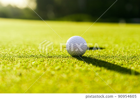 Golf ball on the green grass near hole on a golf course at sunset Golf ball on the green grass near hole on a golf course at sunset 90954605