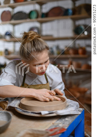 A ceramist makes a plate. Woman in an apron works in a pottery workshop. A ceramist makes a plate. Woman in an apron works in a pottery workshop. 90955273