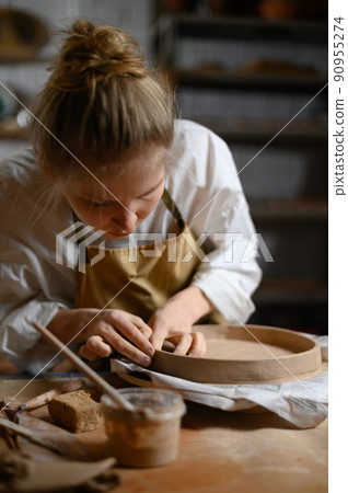 A ceramist makes a plate. Woman in an apron works in a pottery workshop. A ceramist makes a plate. Woman in an apron works in a pottery workshop. 90955274