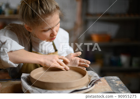 A ceramist makes a plate. Woman in an apron works in a pottery workshop. A ceramist makes a plate. Woman in an apron works in a pottery workshop. 90955275