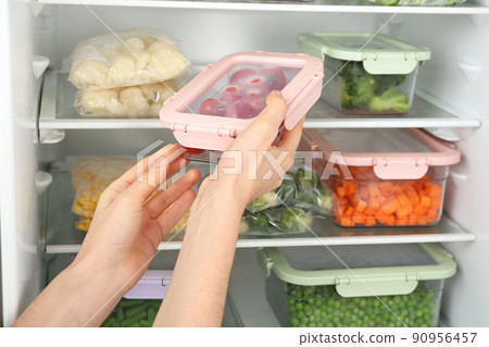 Woman putting container with tomatoes in refrigerator with frozen vegetables, closeup 90956457