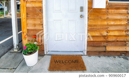 Panorama White front door decorated with flowers on a pot and doormat with welcome sign Panorama White front door decorated with flowers on a pot and doormat with welcome sign 90960017
