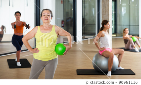 Portrait of mature woman standing with fitness ball during group training at gym Portrait of mature woman standing with fitness ball during group training at gym 90961116