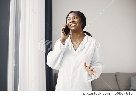 Young female black doctor standing in her office at clinic near coach. Woman wearing white medical coat. Brunette medical worker using a phone. Young female black doctor standing in her office at clinic near coach. Woman wearing white medical coat. Brunette medical worker using a phone. 90964616