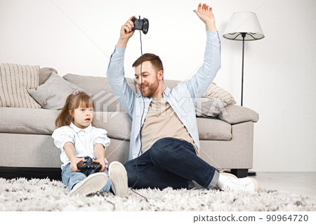 Loving father playing with his daughter with Down syndrome at home together. Man and girl sitting on a floor near sofa and holding joysticks. Bearded man wearing blue shirt and girl white shirt. Loving father playing with his daughter with Down syndrome at home together. Man and girl sitting on a floor near sofa and holding joysticks. Bearded man wearing blue shirt and girl white shirt. 90964720