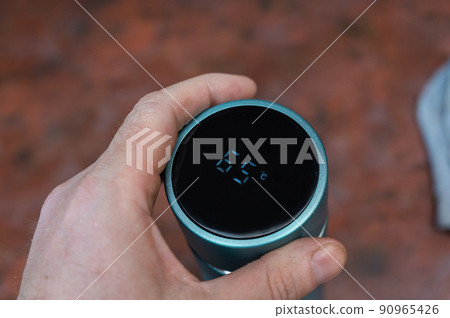 A European man holds a closed thermos against a red background. Turquoise vacuum flask. Lid with LED digital display. Temperature 65 degrees Celsius. Inside the room. Selective Focus. Series part. A European man holds a closed thermos against a red background. Turquoise vacuum flask. Lid with LED digital display. Temperature 65 degrees Celsius. Inside the room. Selective Focus. Series part. 90965426