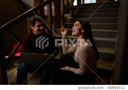Friendly team of a pretty woman and handsome confident mature man works together, sitting on stairs in creative office and discussing plans, projects Friendly team of a pretty woman and handsome confident mature man works together, sitting on stairs in creative office and discussing plans, projects 90969312