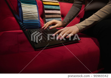 Close-up. Hands of a woman working on laptop, sitting on a red sofa with upholstery fabric samples on background. New home interior design project 90969342