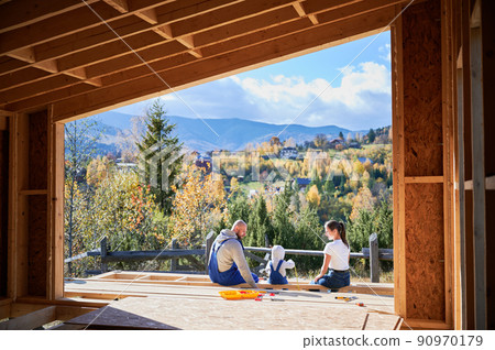 Father, mother and son building wooden frame house. Toddler boy helping his daddy, while woman looking for them on construction site. View from inside a house. Carpentry, family concept. 90970179