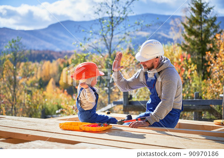 Father with toddler son building wooden frame house. Male builder giving high five to kid on construction site, wearing helmet and blue overalls on sunny day. Carpentry and family concept. 90970186